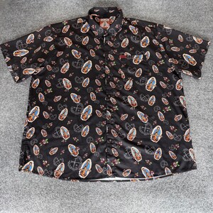 May include: Black short-sleeve button-down shirt with an all-over pattern of religious imagery, including the Virgin Mary, roses, and crosses. The shirt has a classic collar, a chest pocket, and the brand name "DIXXON" on the collar.