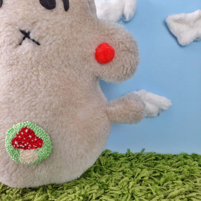 May include: A plush toy shaped like a gray monster with a red pom-pom on its cheek and a green embroidered mushroom on its side. The toy is sitting on a green carpet in front of a blue background.