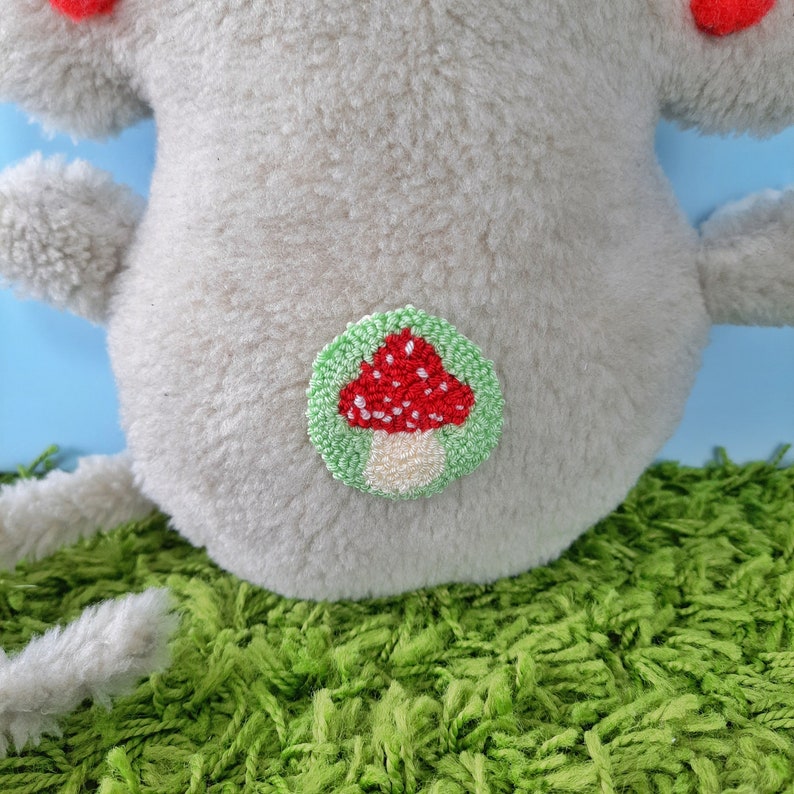 May include: A plush gray stuffed animal with a red and white mushroom embroidered on its back. The mushroom is surrounded by a green circle.