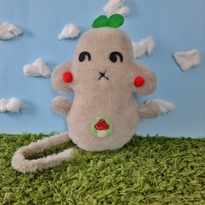 May include: A plush toy shaped like a gray monster with a green clover on its head. It has red cheeks, black eyes, and a mushroom embroidered on its chest. The toy is sitting on a green carpet.