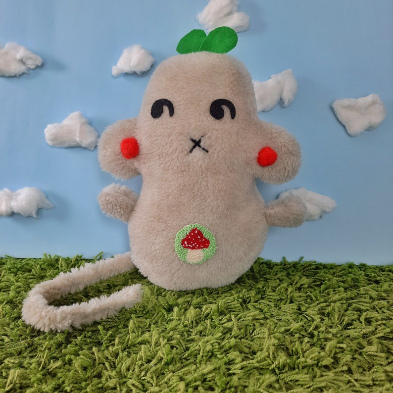 May include: A plush toy shaped like a gray monster with a green clover on its head. It has red cheeks, black eyes, and a mushroom embroidered on its chest. The toy is sitting on a green carpet.