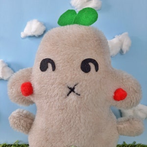 May include: A plush toy shaped like a cloud with a green leaf on top. The toy has a beige fur, black eyes, and red cheeks.