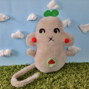 May include: A plush toy shaped like a gray mouse with a green clover on its head and a red mushroom on its belly. The toy is sitting on a green grassy surface with white cotton clouds in the background.