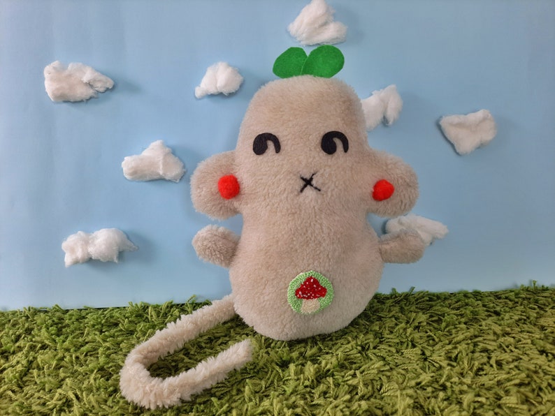 May include: A plush toy shaped like a gray mouse with a green clover on its head and a red mushroom on its belly. The toy is sitting on a green grassy surface with white cotton clouds in the background.
