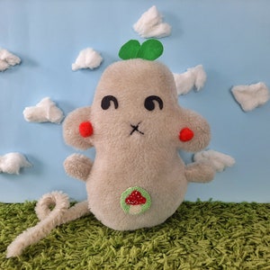 May include: A gray plush toy mouse with red cheeks, a green clover on its head, and a mushroom patch on its belly. The mouse is sitting on a green carpet in front of a blue background with white cotton clouds.