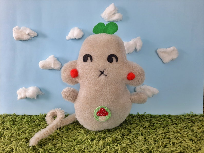 May include: A gray plush toy mouse with red cheeks, a green clover on its head, and a mushroom patch on its belly. The mouse is sitting on a green carpet in front of a blue background with white cotton clouds.