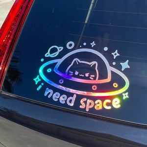 May include: A black car with a rainbow iridescent sticker on the rear window. The sticker features a cat inside a UFO with the text "I need space".