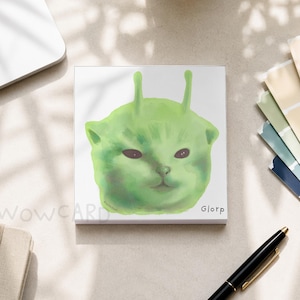 May include: A square notepad featuring a green, alien-like cat illustration with two antennae on its head. The word "Glorp" is printed in the bottom right corner, and "WOWCARD" is faintly visible in the bottom left corner of the white paper.