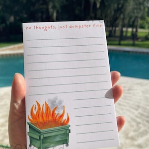 May include: A white rectangular notepad with horizontal gray lines. The top features the red text "no thoughts, just dumpster fire". The bottom left corner displays a watercolor illustration of a green dumpster with orange and yellow flames.