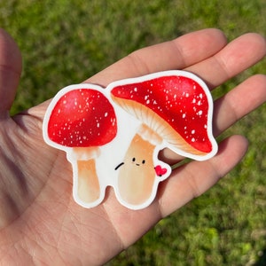 May include: Two red and white mushroom stickers with white spots. The larger mushroom has a smiling face and is holding a small red heart.