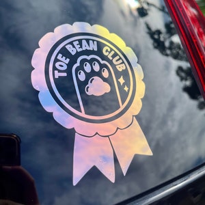 May include: A holographic sticker with a paw print in the center and the text "Toe Bean Club" around the paw print. The sticker is in the shape of a ribbon.
