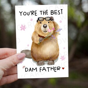 May include: A greeting card featuring a cartoon beaver wearing glasses and holding flowers. The card reads "You're the Best Dam Father" in black text with pink floral accents. The beaver is brown with a large tail.