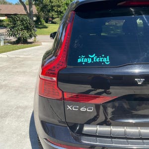 May include: Black SUV with a teal "stay feral" sticker on the back window. The car has a silver "XC60" emblem on the back.