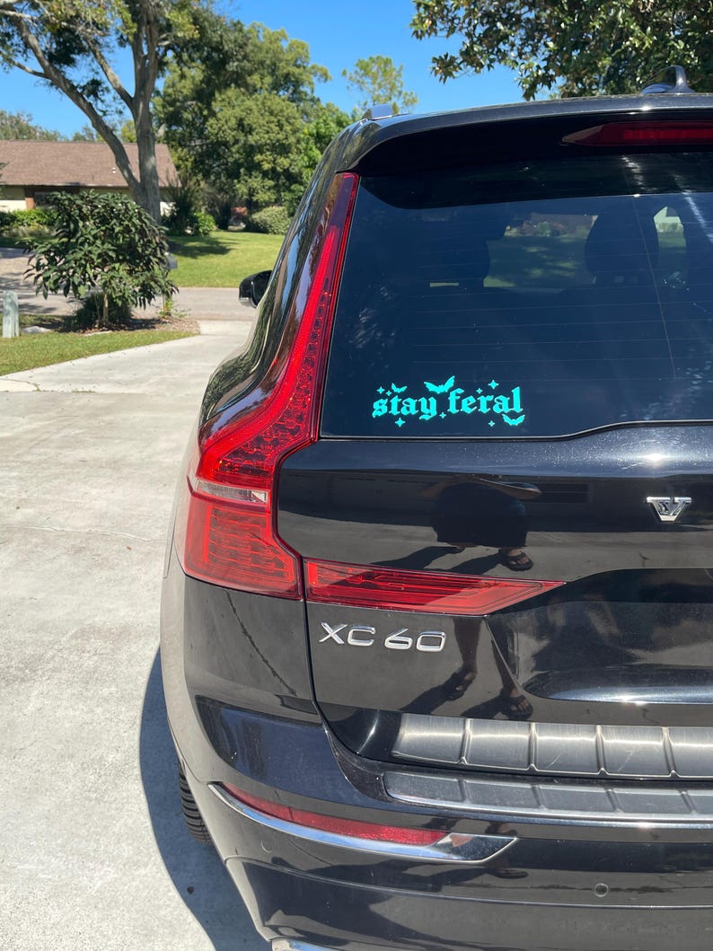 May include: Black SUV with a teal "stay feral" sticker on the back window. The car has a silver "XC60" emblem on the back.