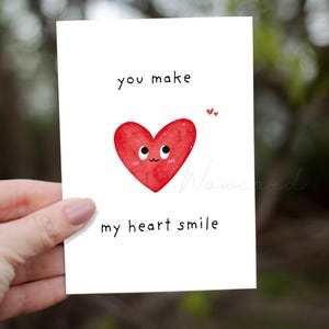 May include: A white greeting card with the text "you make my heart smile" and a red heart illustration with a smiling face. Two small red hearts are above the large heart.