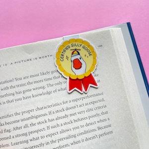 May include: A yellow and red bookmark with a cartoon goose wearing a pilot hat and a red ribbon. The bookmark has the text "Certified Silly Goose" on it.