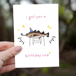 May include: A white greeting card with a whimsical illustration. A fish wearing a party hat and holding a party horn is being held up by a cat. The text reads "I got you a birthday cod".