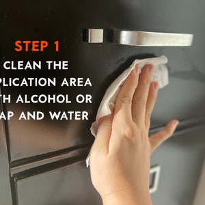 Puede incluir: Un caj&oacute;n de armario negro se limpia con un pa&ntilde;o blanco. El texto "STEP 1 CLEAN THE APPLICATION AREA WITH ALCOHOL OR SOAP AND WATER" se muestra en naranja y blanco en el armario.