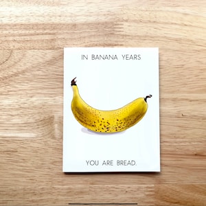 May include: A white card with the text "In Banana Years You Are Bread."  A cartoon illustration of a yellow banana with brown spots is in the center of the card.