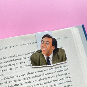 May include: A bookmark featuring a cartoon illustration of Nicolas Cage with a surprised expression. The bookmark is white with a light blue background and the text "ARS 195" in the top right corner.