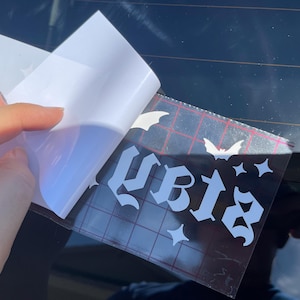 May include: A white vinyl sticker with the word "slay" in gothic lettering, with bats and stars around it. The sticker is being applied to a car window.