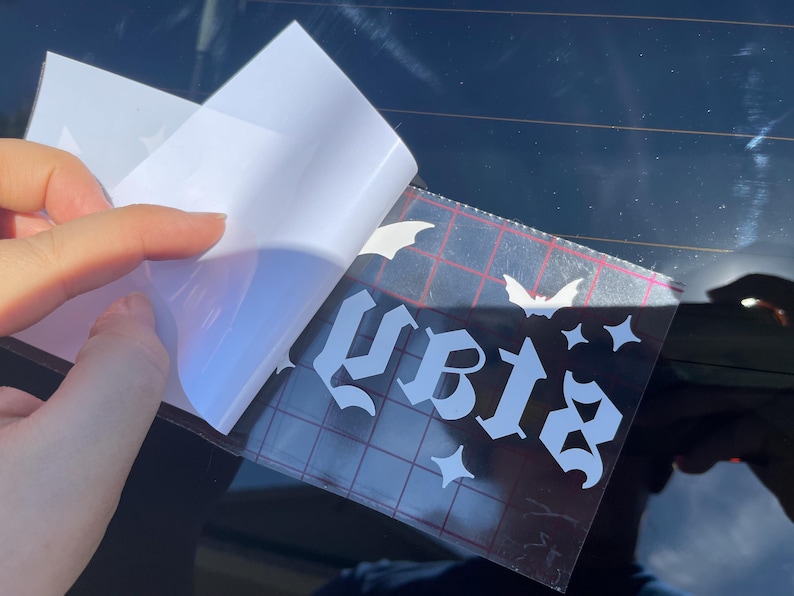 May include: A white vinyl sticker with the word "slay" in gothic lettering, with bats and stars around it. The sticker is being applied to a car window.