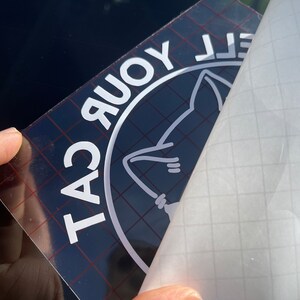 K&ouml;nnte beinhalten: Ein wei&szlig;er Vinyl-Aufkleber mit einem schwarzen Kreis um den Text "TAC YOUR CAT" und einer wei&szlig;en Silhouette einer Katze im Kreis. Der Aufkleber wird von einer transparenten Transferfolie abgezogen.