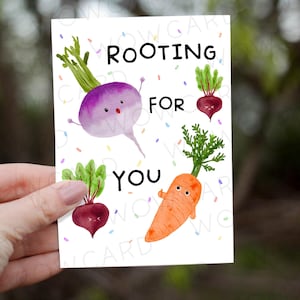 May include: A greeting card with the text "Rooting for You" and illustrations of cartoon vegetables. The vegetables include a purple turnip, a red beet, and an orange carrot. The card is held in a person's hand.