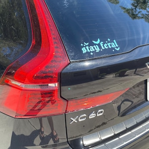 May include: A black SUV with a "stay feral" sticker on the back window. The sticker is in a light blue color with a gothic font and features bats and stars. The car has a silver "XC60" emblem on the back.
