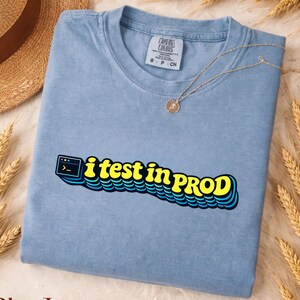 May include: A blue jean coloured t-shirt featuring a graphic print with a computer terminal icon and the yellow text "i test in PROD" with a blue shadow effect. The shirt has a crew neckline and a tag indicating size small, petite, or child.