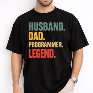 May include: A black short-sleeved t-shirt featuring the text HUSBAND. DAD. PROGRAMMER. LEGEND. printed in a stacked layout. The word HUSBAND is in teal, DAD is in yellow, PROGRAMMER is in tan, and LEGEND is in red, all with a distressed texture.
