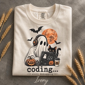 May include: An ivory t-shirt featuring a graphic of a ghost wearing headphones, sitting at a laptop with a black cat and a pumpkin. The design includes a full moon, bats, and a coffee cup. Text reads: coding... still debugging.
