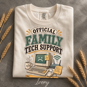 Op de afbeelding: Een ivoorkleurig t-shirt met een opdruk "OFFICIAL FAMILY TECH SUPPORT" boven een illustratie van een vintage computer met een verdrietig gezicht op het scherm, een toetsenbord, een muis, een koffiemok en een wifi-router.