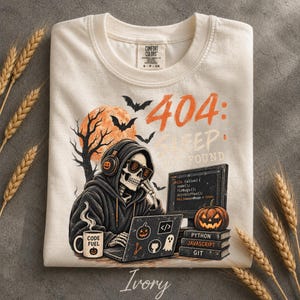 May include: An ivory t-shirt featuring a graphic of a skeleton wearing a hoodie and headphones while working on a laptop. The design includes a full moon, bats, a pumpkin, and text reading 404: Sleep Not Found, Python, Javascript, and Git.