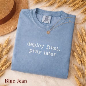 Puede incluir: Una camiseta azul vaquero con el texto "deploy first, pray later" en blanco. Un collar dorado con un colgante descansa sobre la camiseta. Un sombrero de paja y espigas de trigo están en el fondo.