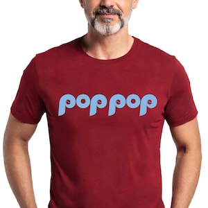 以下が含まれることがあります： 明るい青色の「poppop」という文字が特徴的な、スタイリッシュなフォントのマルーン色のTシャツ。 短い袖のシャツは、灰色のひげと短い髪の人が着ています。