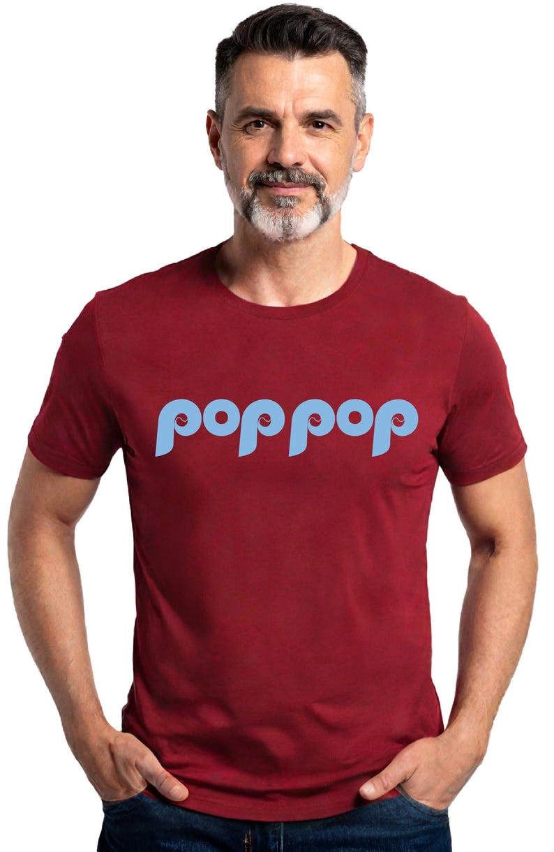 以下が含まれることがあります： 明るい青色の「poppop」という文字が特徴的な、スタイリッシュなフォントのマルーン色のTシャツ。 短い袖のシャツは、灰色のひげと短い髪の人が着ています。