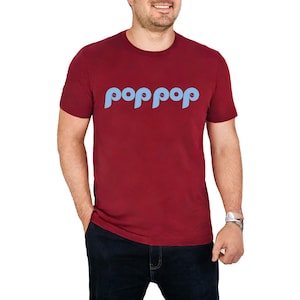 以下が含まれることがあります： ライトブルーの「poppop」という文字が丸みを帯びたフォントでプリントされた、マルーン色のTシャツ。半袖のシャツは人が着ています。その人は濃い青色のジーンズとシルバーの腕時計をしています。