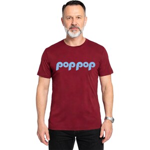 以下が含まれることがあります： 明るい青色の「poppop」という文字がカーブしたデザインの、マルーン色のクルーネックTシャツ。半袖で、人が着ています。その人はシルバーの腕時計と黒いズボンを着用しています。