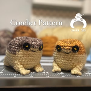 Könnte beinhalten: Zwei gehäkelte Frosch-Plüschtiere, eines braun und beige, das andere orange und beige, mit schwarzen Knopfaugen. Die Plüschtiere sitzen auf einer grauen Oberfläche mit einem Lineal.
