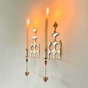 May include: Two brass wall sconces with lit candles. The sconces have a decorative, openwork design with a series of circles and a pointed top. The candles are ivory-colored and have a warm glow.