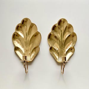 May include: A pair of gold-colored wall sconces. Each sconce has a leaf-like design with a candle holder and a loop for hanging. The sconces are made of metal and have a shiny finish. The overall design is elegant and decorative.