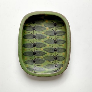 May include: A rectangular ceramic tray with rounded corners, featuring a matte green finish. The interior surface is decorated with a repeating botanical pattern of dark grey leaves and small yellow dots arranged on thin black vertical stems.