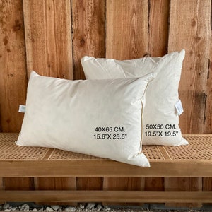 Könnte beinhalten: Zwei cremefarbene Kissen, eines rechteckig (40x65 cm) und eines quadratisch (50x50 cm). Die Kissen werden auf einer gewebten Bank vor einer Holzwand präsentiert. Die Abmessungen in Zoll sind ebenfalls angegeben.