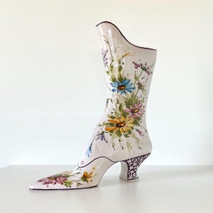 Puede incluir: Un jarrón de cerámica blanca con forma de bota, con punta afilada y diseño floral. El jarrón presenta un tacón alto con un diseño estampado y está adornado con flores coloridas en tonos azules, amarillos y rosas.
