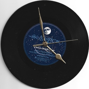 Peut inclure: Une horloge circulaire faite à partir d'un disque vinyle noir, avec des aiguilles dorées. L'étiquette centrale est bleue avec du texte blanc et un motif de lune. Le texte sur l'étiquette indique "The Mission" et "Deliverance".