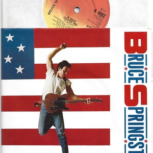 May include: A red, white and blue American flag with a man in blue jeans and a white t-shirt jumping in front of it while playing a guitar. The text "BRUCE SPRINGSTEEN" is on the right side of the image. The text "A 4662" and "INCLUDES A FREE POSTER" are at the bottom of the image.