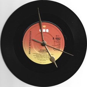 Puede incluir: Un reloj de vinilo negro con una etiqueta roja y amarilla con el texto "Bruce Springsteen - A Side Cover Me" y "A 4662" impreso en él. Las manecillas del reloj son doradas.