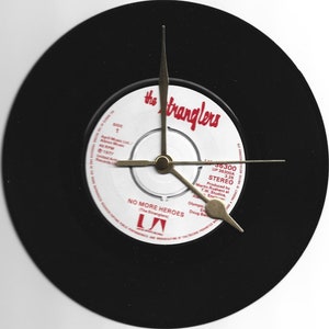 Puede incluir: Un disco de vinilo negro con una etiqueta blanca. La etiqueta tiene el texto "the Stranglers" en letras rojas y "NO MORE HEROES (The Stranglers)" en letras negras. El disco es un disco de 45 RPM y tiene el título de la canción "No More Heroes" en él. El disco es un producto de Arista Records Ltd.