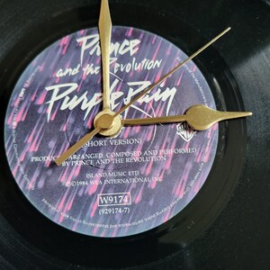 Pode incluir: Um relógio de vinil preto com a arte do álbum "Purple Rain" de Prince and the Revolution. Os ponteiros do relógio são dourados. O título do álbum e o nome do artista são visíveis no rótulo roxo e branco.